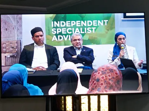 Rumana Mahomed speaking into a microphone beside two other panellists, under an Independent Specialist Advisor slide